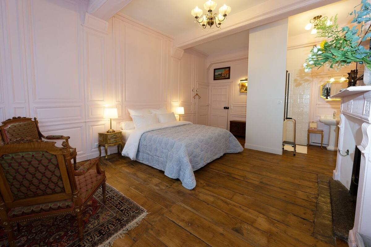 A bedroom with a bed, nightstands, a chair, a fireplace, and a mirror.