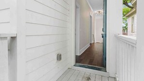 Eine weiße Außenwand mit horizontaler Schalung, eine Holzterrasse und eine Glastür, die zu einem Innenflur führt.