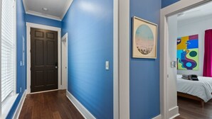 A hallway with blue walls, a wooden floor, a dark door, and a bed with a colorful painting and pink curtains in the adjacent room. - Fun & Renovated Artsy Downtown Apt Dog Friendly with Fully Fenced Backyard and Private Parking (Savannah)