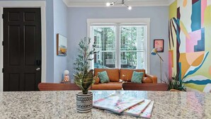 A living room with a brown sofa, a coffee table with magazines, a potted plant, and a window with blinds. - Fun & Renovated Artsy Downtown Apt Dog Friendly with Fully Fenced Backyard and Private Parking (Savannah)