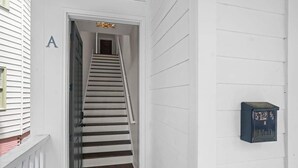 Fachada blanca con una escalera de madera que conduce a una puerta marcada con la letra 'A'. Un buzón está adosado a la pared.