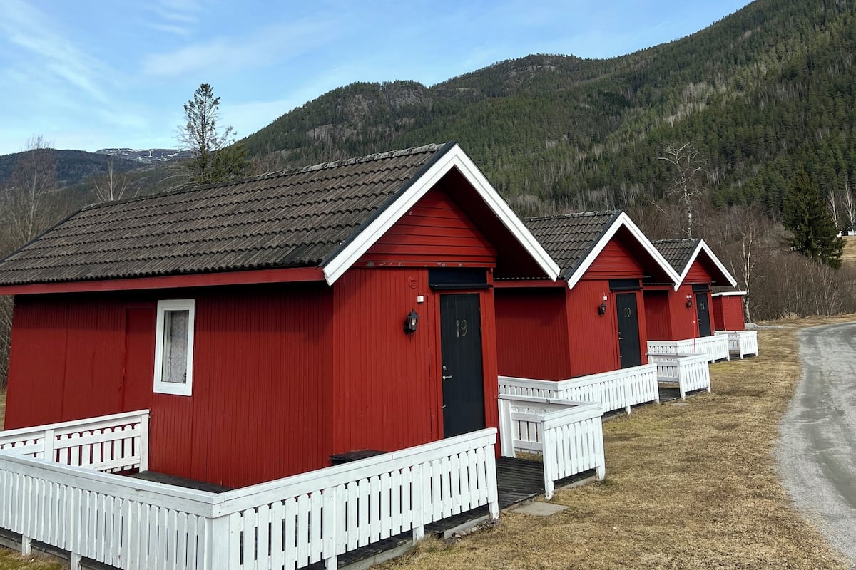 En rad röda stugor med vita räcken, numrerade från 19, som står i kontrast till en klarblå himmel och en skogbeklädd sluttning i bakgrunden.