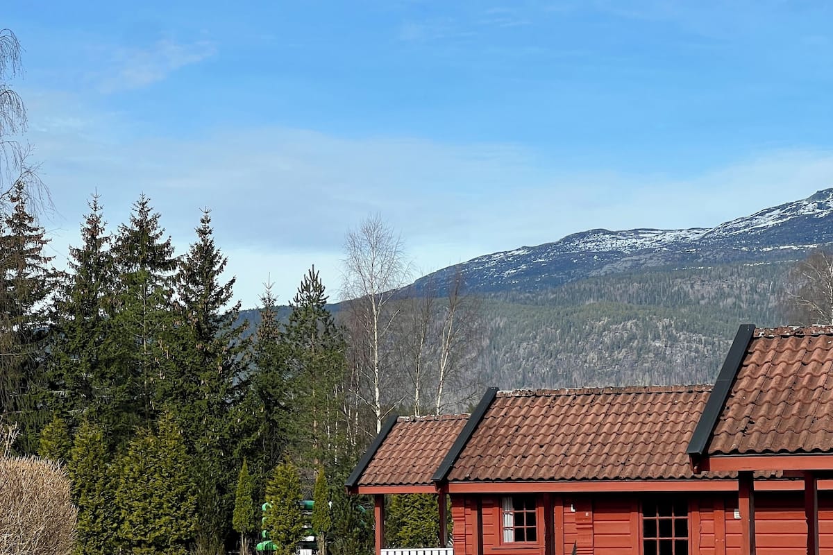En röd stuga med en grusuppfart och ett vitt staket, omgiven av träd och berg.