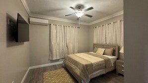 Un dormitorio con cama, mesita de noche, lámpara, ventilador de techo y televisor en la pared.