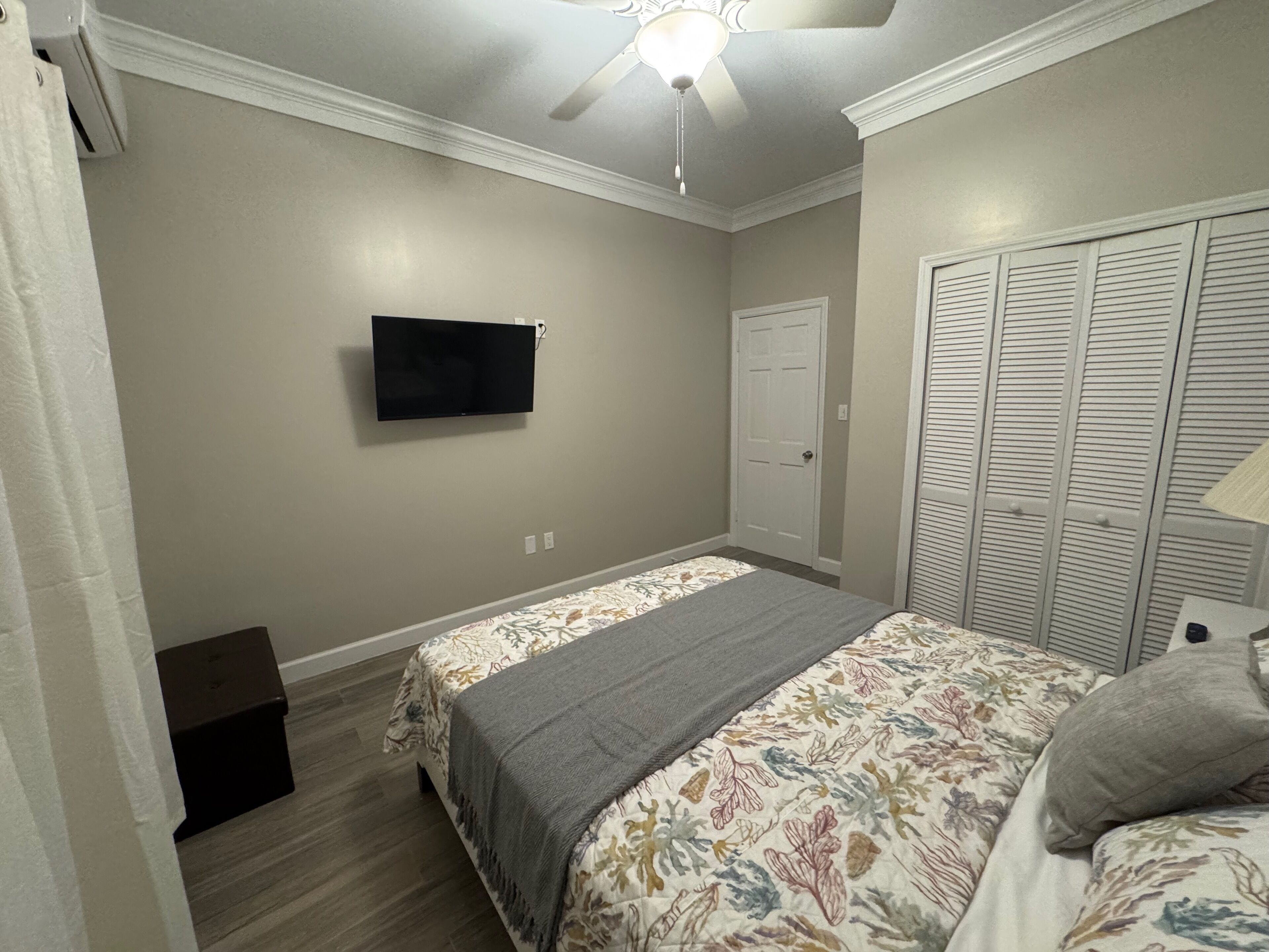 Une chambre à coucher comprenant un lit, un téléviseur fixé au mur, une armoire avec des portes à persiennes et un ventilateur de plafond.