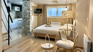 Ein modernes Hotelzimmer mit einem Bett, einem Korbstuhl, einem kleinen Tisch, einem Fernseher und einer Küchenzeile mit einer Steinwand.