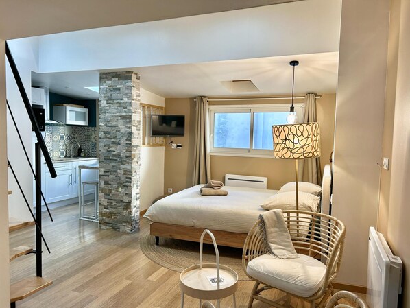A modern hotel room with a bed, a wicker chair, a small table, a TV, and a kitchen area with a stone wall.