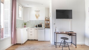 Eine kompakte Küche mit weißen Schränken, einer Mikrowelle, einem kleinen Kühlschrank, einer Spüle und einem Holztisch mit einem Hocker. Ein Flachbildfernseher ist an der Wand über dem Tisch angebracht.