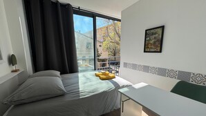 A bedroom with a bed, a desk, a window with curtains, a small plant, and a framed picture on the wall. - L'Amandier - Lovely New 2-Bedroom apt in Aix City Center (Aix-en-Provence)