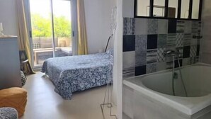 A bedroom with a bed, a window, and a view of greenery outside. There is a bathroom with a bathtub and a shower.