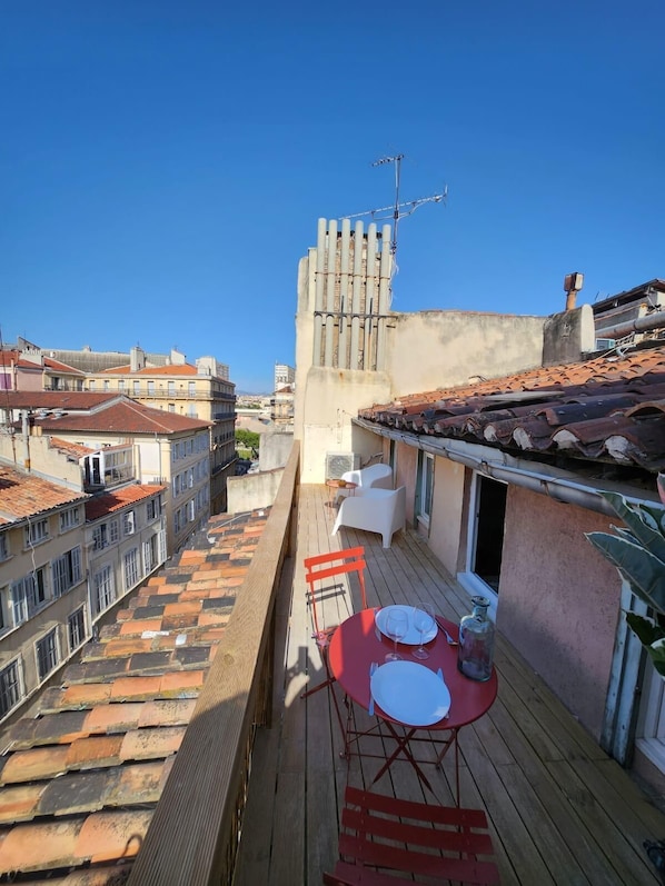 A rooftop terrace with a red table set for dining, white chairs, and a view of the cityscape.