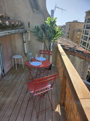 Una terraza de madera con una mesa y sillas de metal rojo, una planta en maceta y un taburete blanco pequeño.