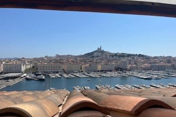 Un paisaje urbano costero con una estructura destacada en una colina, un puerto deportivo con numerosos barcos y edificios en primer plano.