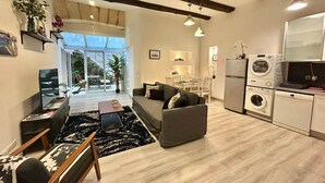 Ein geräumiger Wohnbereich mit Sofa, Couchtisch und Esstisch. Es gibt eine Küchenzeile mit Geräten und ein großes Fenster.