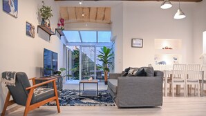 Ein modernes Wohnzimmer mit grauem Sofa, Holzschaukelstuhl und einem kleinen Tisch. Es befinden sich Blumentöpfe, ein großes Fenster und ein Essbereich mit weißen Stühlen dort.