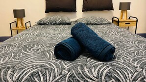 A neatly made bed with a patterned bedspread, two pillows, and rolled towels. There are two bedside tables with lamps and a small wooden chair. - Chez Florence - Spacious 1BR in Historic City Center (Aix-en-Provence)