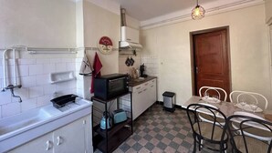 A compact kitchen with white cabinets, a microwave, a toaster, and a sink. There's a dining area with a table and chairs. - Chez Monique - 1BR in historic city center, mairie square (Aix-en-Provence)