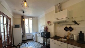 A compact kitchen with a washing machine, microwave, and a coffee maker. There is a dining table with chairs and a checkered floor. - Chez Monique - 1BR in historic city center, mairie square (Aix-en-Provence)