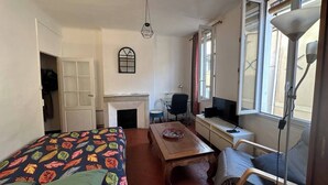 A bedroom with a bed, a desk, a chair, a small table, a fireplace, and a window with curtains. - Chez Monique - 1BR in historic city center, mairie square (Aix-en-Provence)