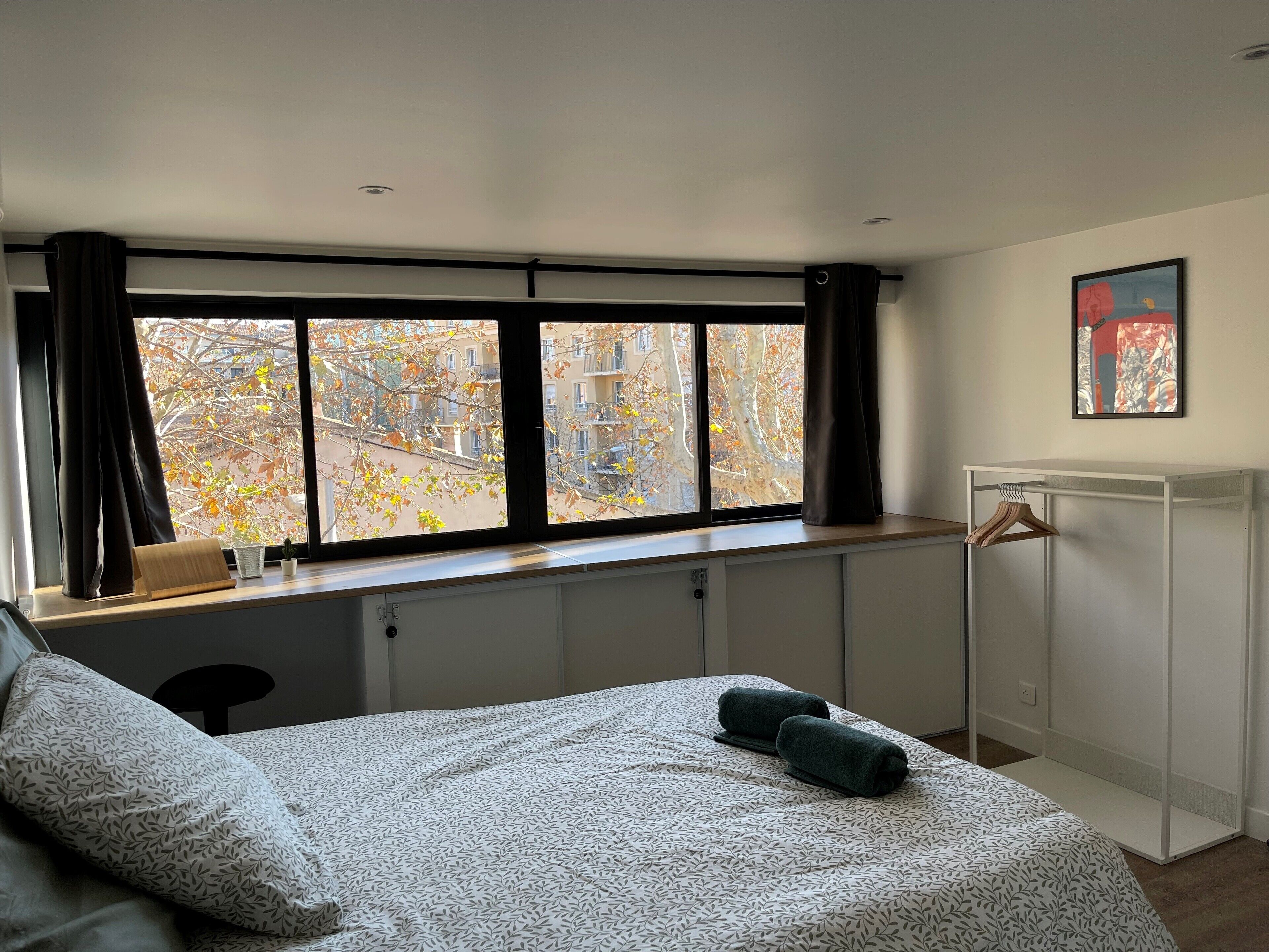 A bedroom with a bed, a window with curtains, a radiator, a coat rack, and a painting on the wall.