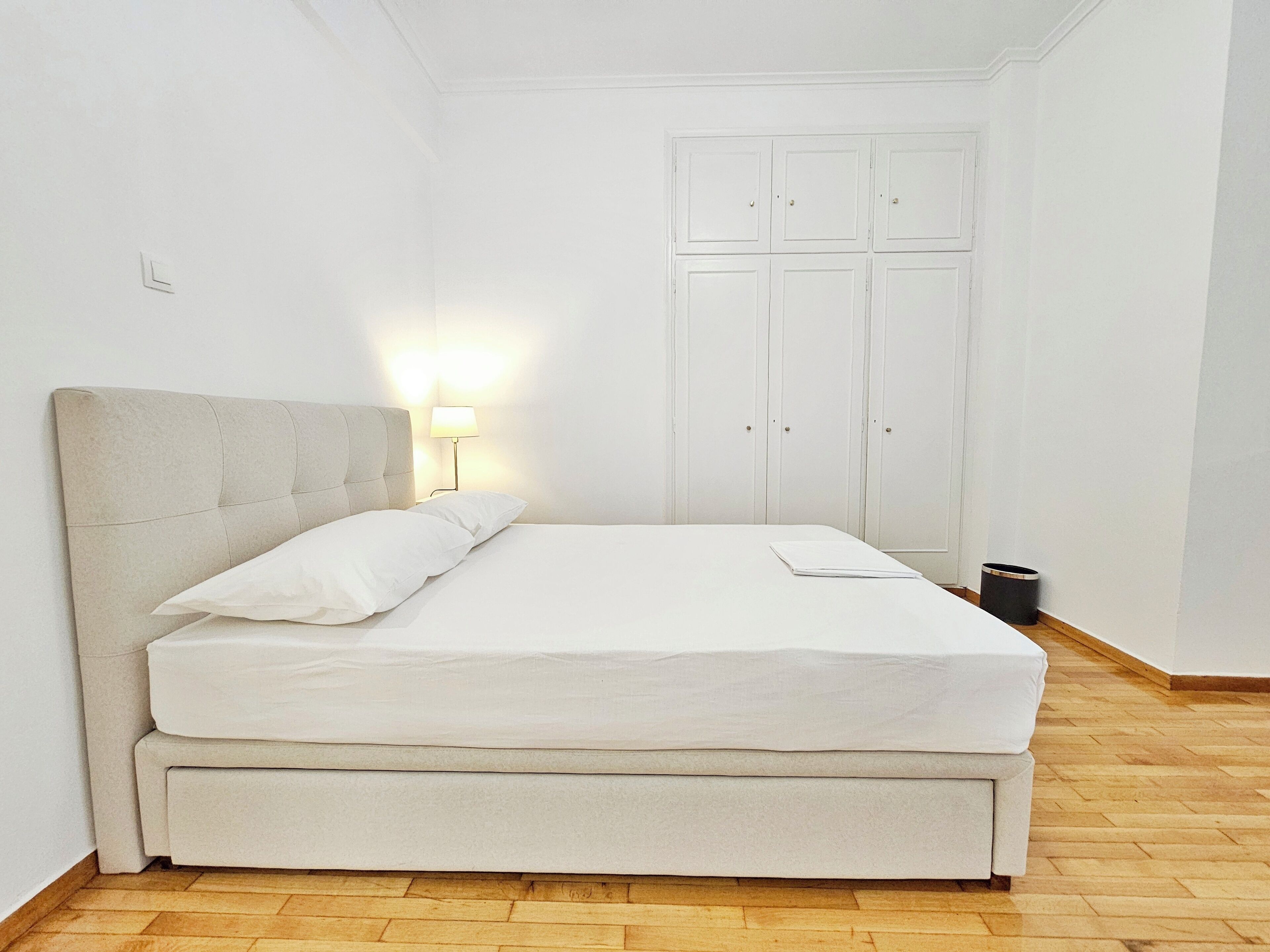 A neatly made bed with a headboard, a bedside lamp, and a small trash bin in a room with white walls and wooden flooring.
