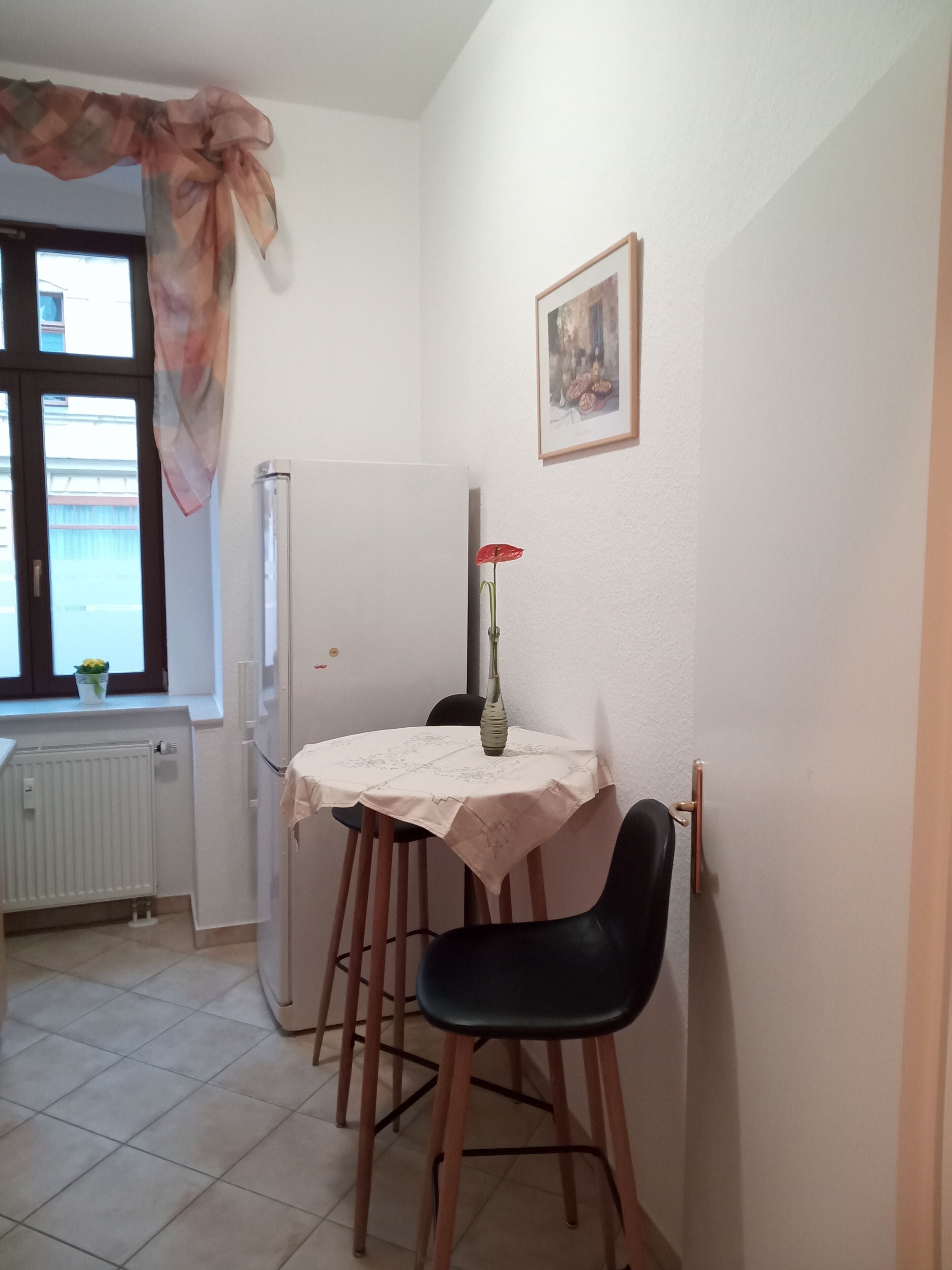 Een knus eetgedeelte met een tafel en stoelen, een radiator, een raam met gordijn en een lijst met een afbeelding aan de muur.
