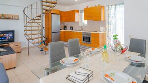 Une cuisine moderne avec des armoires en bois, un four encastré et un réfrigérateur en acier inoxydable. Une table à manger avec des chaises est dressée pour le repas, et un escalier en colimaçon est visible au fond.