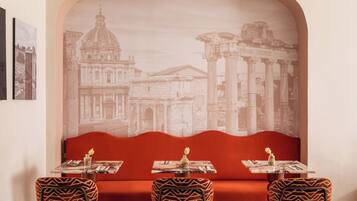 Un ristorante con una zona salotto con panche imbottite rosse, tavoli apparecchiati e un grande murale raffigurante architettura classica.