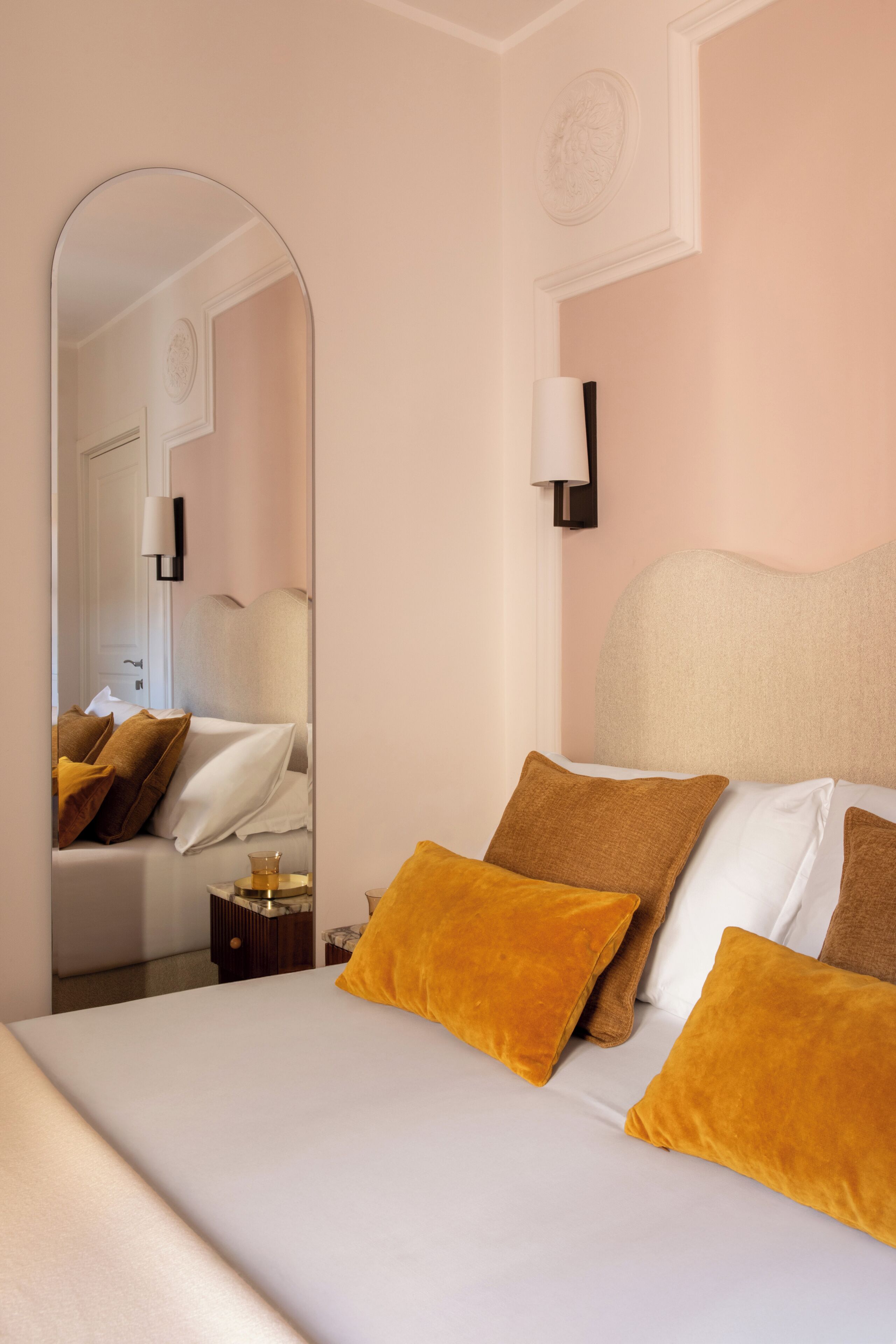 A hotel room with a bed, a nightstand, a mirror, and a wall-mounted lamp.
