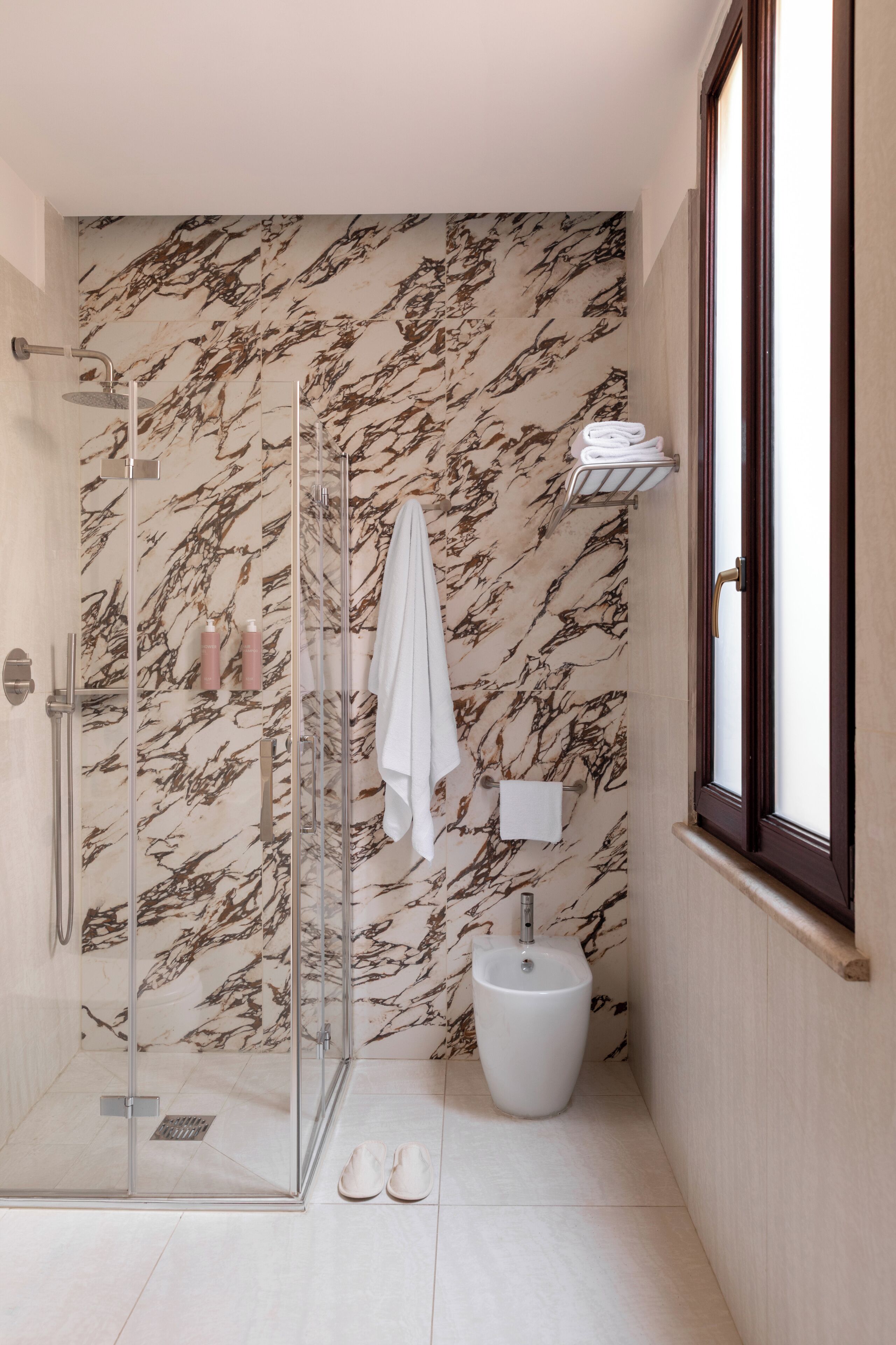 A bathroom with a glass shower enclosure, a marble-patterned wall, a white sink, and a window.