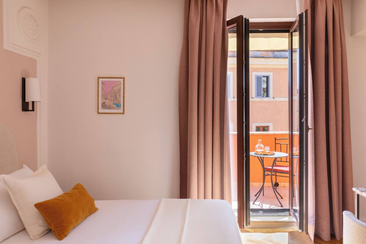 A hotel room with a bed, a window with curtains, a small table, and a chair outside.