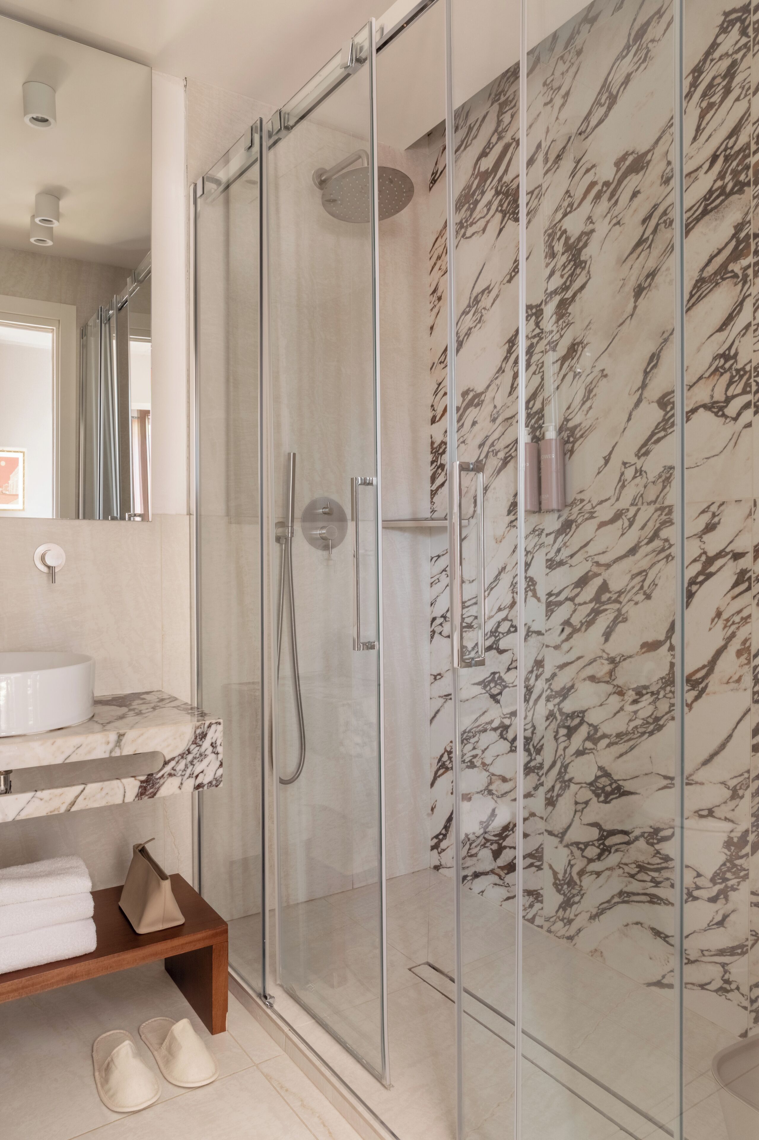 Ein modernes Badezimmer mit einer gläsernen Duschkabine, Marmorwänden, einem weißen Waschbecken und einer Holzbank mit Hausschuhen.