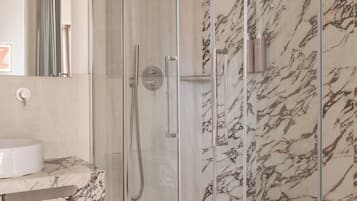 Un baño moderno con ducha envuelta en cristal, paredes de mármol, un lavabo blanco y un banco de madera con zapatillas.