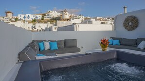 Una terraza en la azotea con un jacuzzi, cojines grises y azules, y vistas a edificios blancos al fondo.