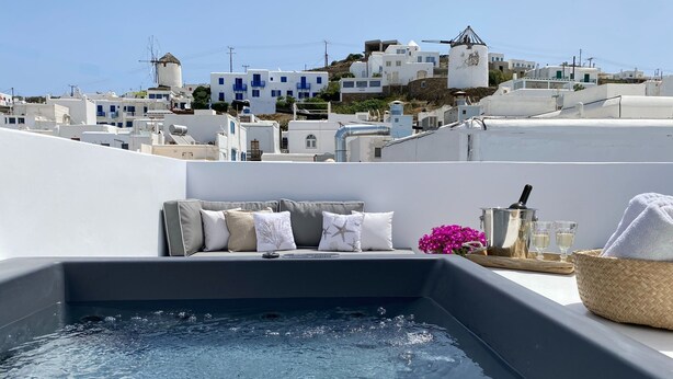 Una piscina en la azotea con vistas a edificios blancos y molinos, un sofá con cojines y una cesta con toallas.
