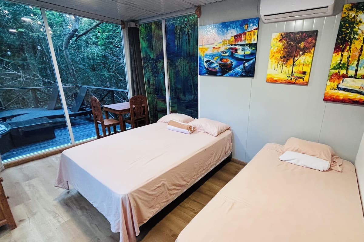 A bedroom with two beds, a wooden table, and chairs. There are large windows with a view of trees and a dock.
