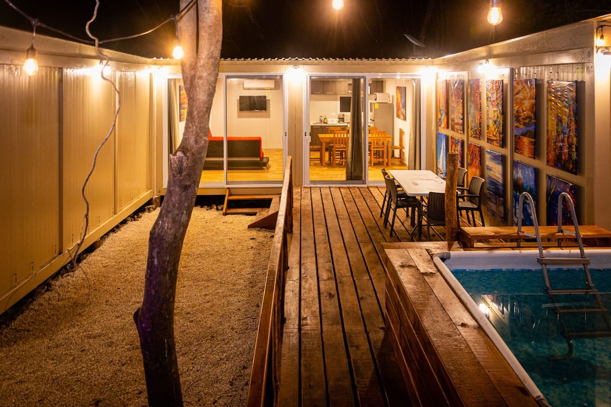A pool area with a wooden deck, a ladder, and a table with chairs. There is a sliding glass door leading to an interior space with a kitchen and dining area.