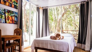A bedroom with a bed, a wooden chair, and a table. There are curtains and a window with a view of trees. - La Ballena Nómada (Akumal)
