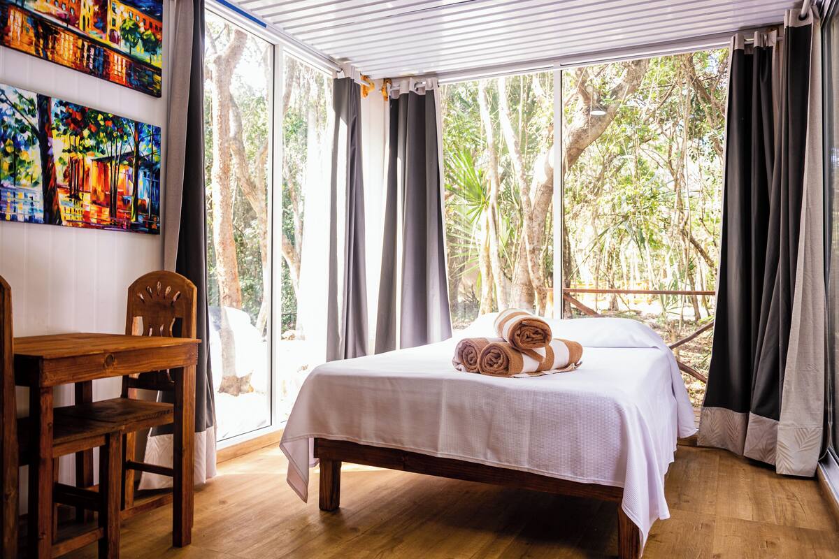 A bedroom with a bed, a wooden chair, and a table. There are curtains and a window with a view of trees.
