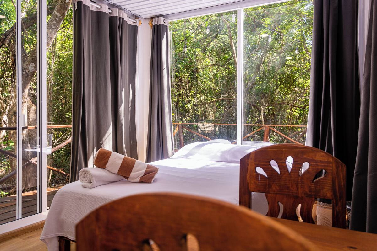 A room with a bed, a wooden chair, and a large window with curtains.