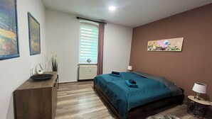 A bedroom with a bed, a dresser, a radiator, a window with blinds, a lamp, and a painting on the wall.