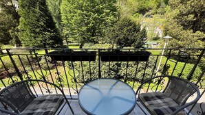 A balcony with a round table and two chairs, overlooking a garden with trees and a house in the background.
