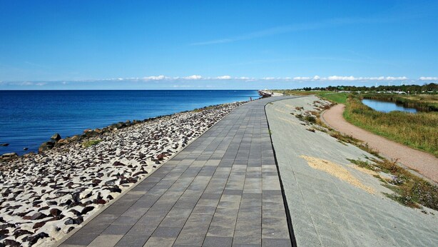 A paved walkway along a coastal area with a clear view of the sea and a small body of water. - Ferienhaus Sturmfrei auf Fehmarn (Fehmarn OT Kopendorf)