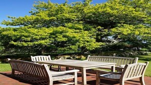 An outdoor patio with a wooden table and benches, surrounded by lush greenery and trees. - Ferienhaus Sturmfrei auf Fehmarn (Fehmarn OT Kopendorf)