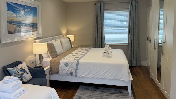 A hotel room with a bed, bedside tables, a nightstand, a chair, a window with curtains, a wall-mounted picture, and a door.