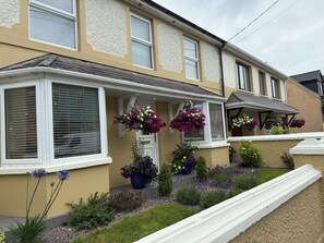 Une maison à deux étages avec un porche à l’avant, des jardinières suspendues et un petit jardin où poussent différentes espèces de plantes.