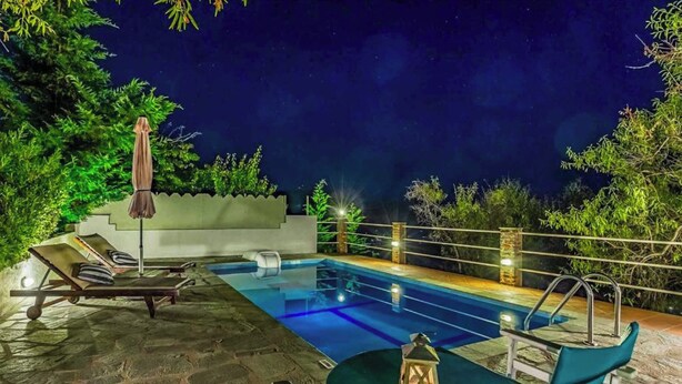 Uma área de piscina ao ar livre à noite, com espreguiçadeiras, uma mesa e um candeeiro.
