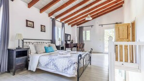 Quarto com teto de madeira, cama com colcha estampada, mesa de cabeceira com abajur e escrivaninha com cadeira.