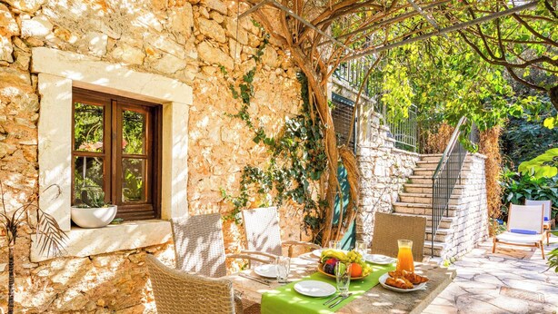 A stone-walled outdoor dining area with wicker chairs, a table set with plates, cutlery, and a fruit bowl. - Stone Cottage - Two Bedroom Villa, Sleeps 4 (Nisaki)