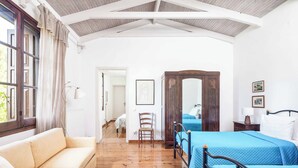 A room with a wooden floor, a sofa, two beds with blue bedding, a wooden wardrobe, and a window with curtains. - Stone Cottage - Two Bedroom Villa, Sleeps 4 (Nisaki)
